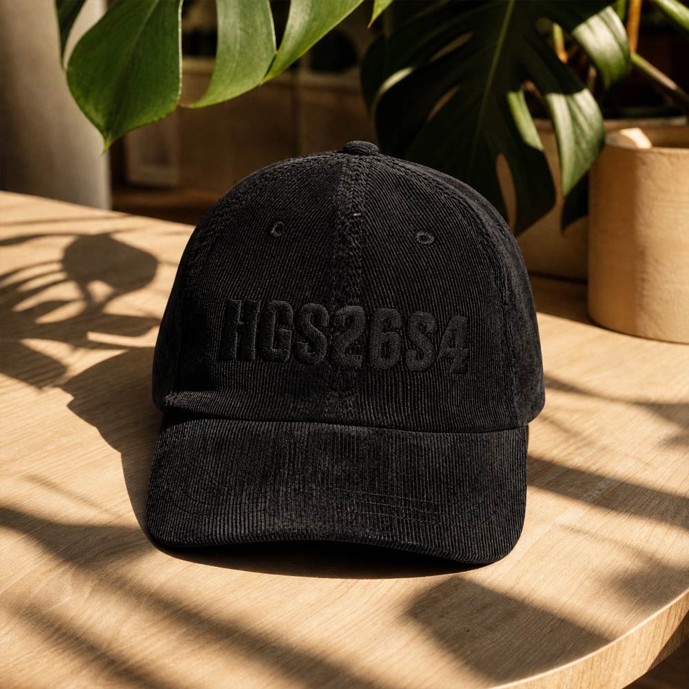 His Good Stuff's Autumn Catalog HGS26S4 Corduroy Cap, front, 100% cotton corduroy with an adjustable golden buckle in the back, on a table. Graphic displays in black thread, 3-D embroidered HGS26S4