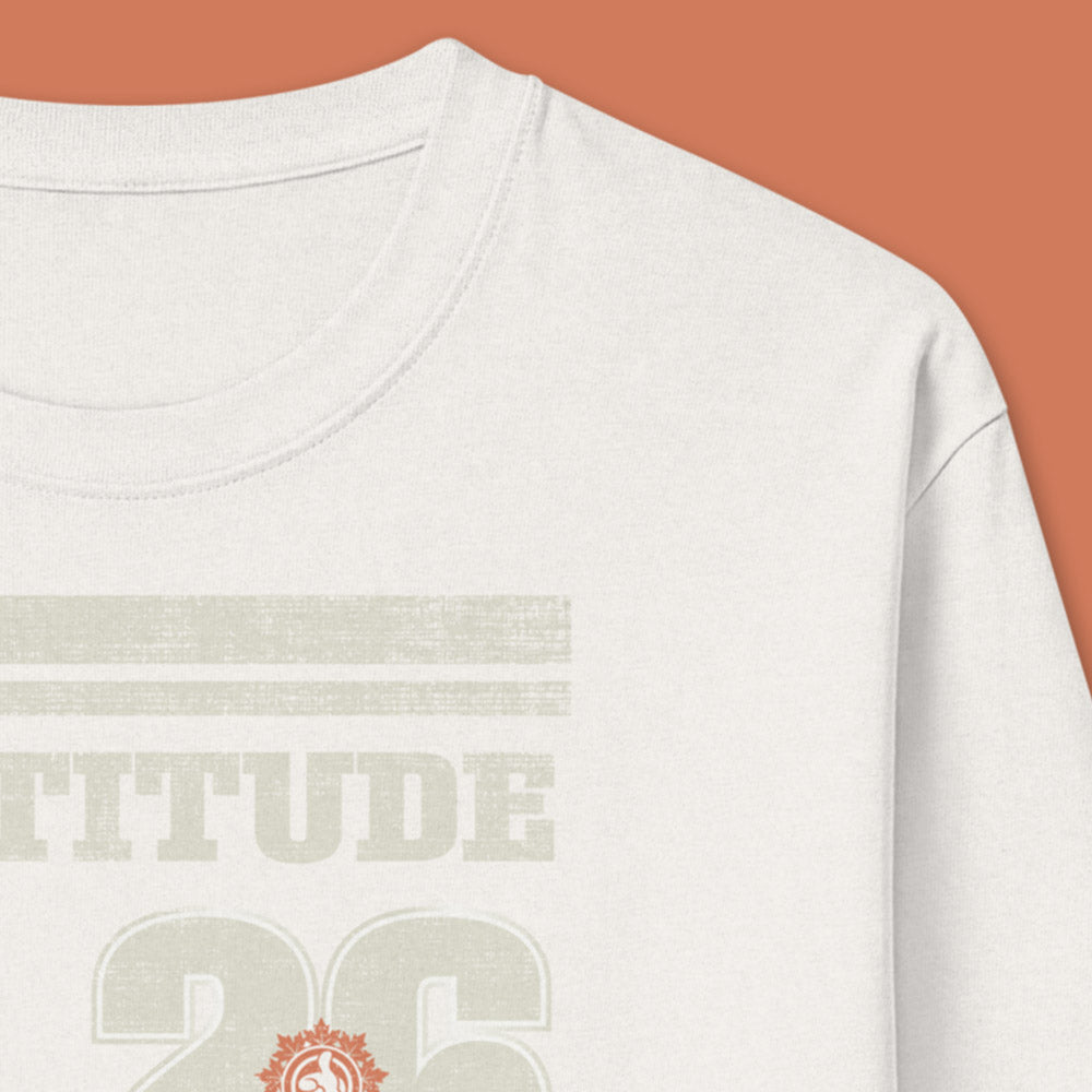 His Good Stuff's Autumn Catalog HGS26S4 Gratitude Jersey, front, heavyweight, 100% ring-spun cotton, white, long-sleeve, detail of fabric against a flat Brandied Melon background. Graphic displays two bars over distressed letters GRATITUDE, a number 26, and the HGS fall thumbs-up icon.
