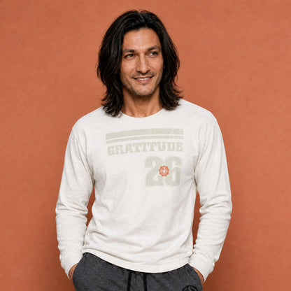 His Good Stuff's Autumn Catalog HGS26S4 Gratitude Jersey, front, heavyweight, 100% ring-spun cotton, white, long-sleeve, on model against a flat Brandied Melon background. Graphic displays two bars over distressed letters GRATITUDE, a number 26, and the HGS fall thumbs-up icon.