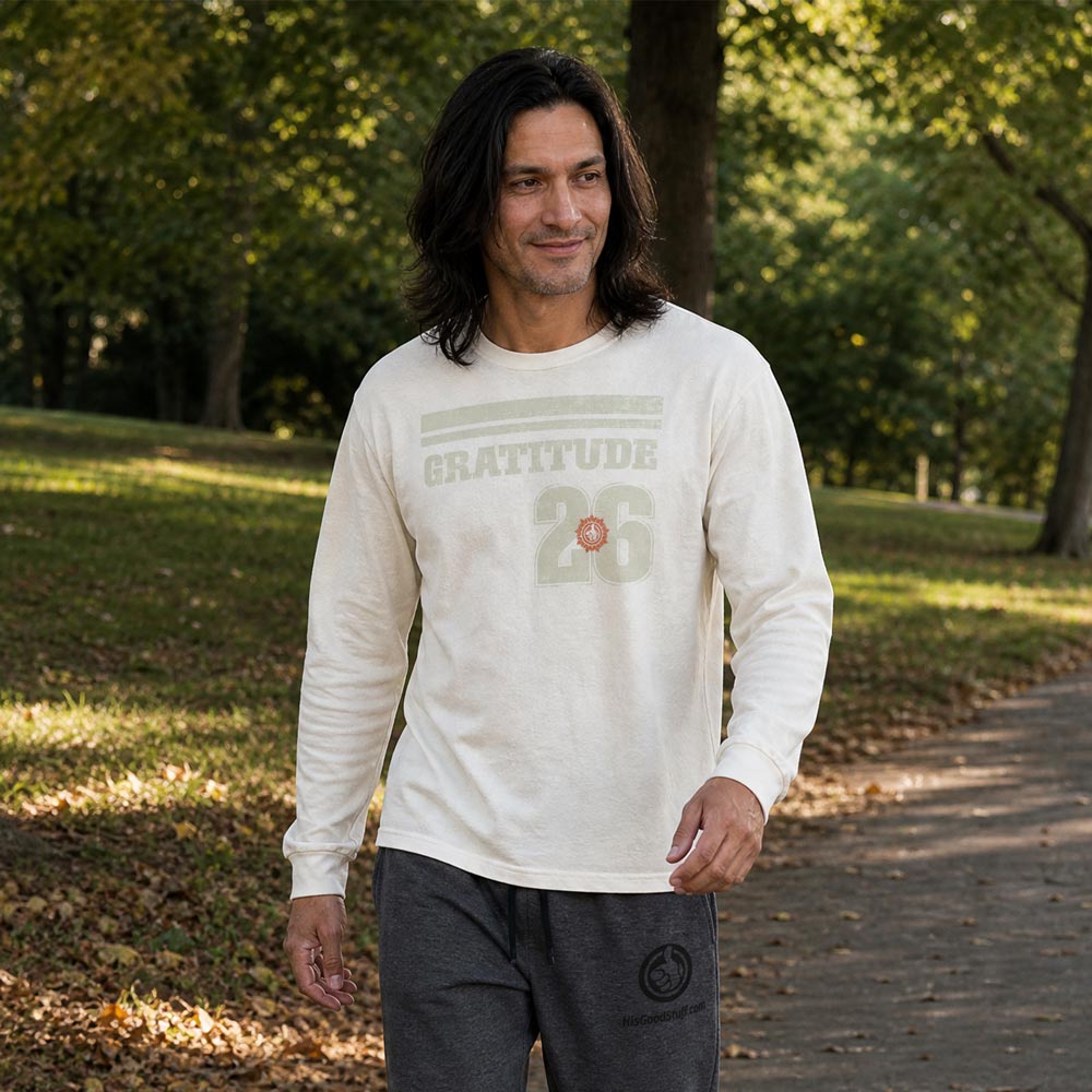 His Good Stuff's Autumn Catalog HGS26S4 Gratitude Jersey, front, heavyweight, 100% ring-spun cotton, white, long-sleeve, on model walking in a city park in the Fall. Graphic displays two bars over distressed letters GRATITUDE, a number 26, and the HGS fall thumbs-up icon.