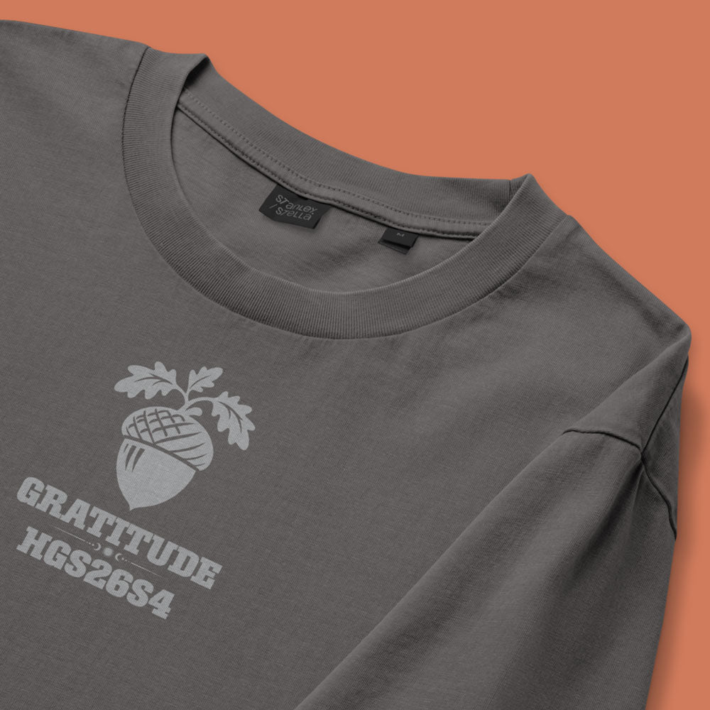 His Good Stuff's Autumn Catalog HGS26S4 Gratitude Tee, front fabric detail, relaxed fit, 100% ring-spun cotton, grey, short-sleeve, front, on a model against a flat Brandied Melon background. Graphic displays an acorn graphic above the letters "GRATITUDE" and "HGS26S4".