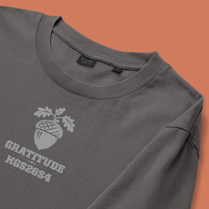 His Good Stuff's Autumn Catalog HGS26S4 Gratitude Tee, front fabric detail, relaxed fit, 100% ring-spun cotton, grey, short-sleeve, front, on a model against a flat Brandied Melon background. Graphic displays an acorn graphic above the letters "GRATITUDE" and "HGS26S4".