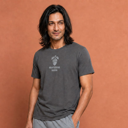 His Good Stuff's Autumn Catalog HGS26S4 Gratitude Tee, closeup front, relaxed fit, 100% ring-spun cotton, grey, short-sleeve, front, on a model against a flat Brandied Melon background. Graphic displays an acorn graphic above the letters "GRATITUDE" and "HGS26S4".