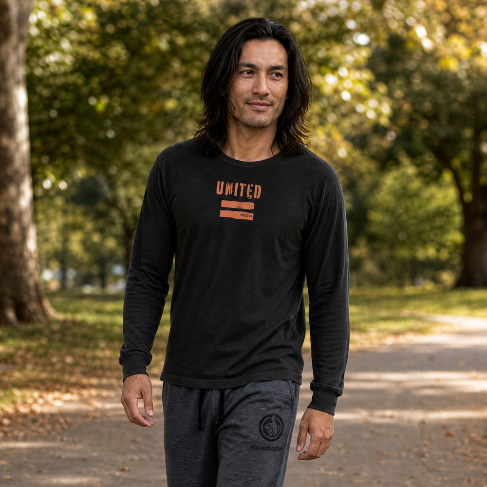 His Good Stuff's Autumn Catalog HGS26S4 United Equality, front, heavyweight, 100% ring-spun cotton, black, long-sleeve, on a model walking in a city park in the Fall. Graphic displays two bars beneath the distressed letters "EQUALITY".
