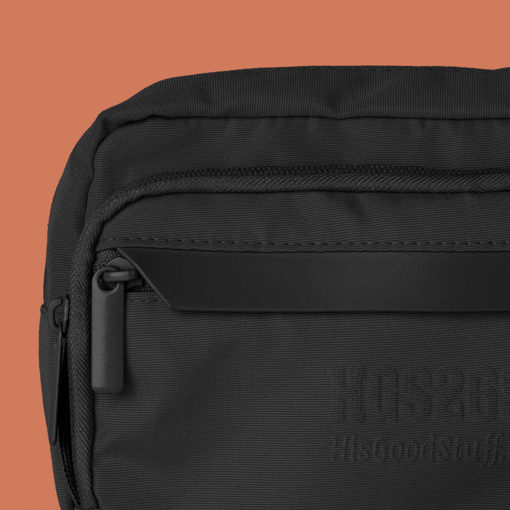 His Good Stuff's Autumn Catalog HGS26S4 Urban Bag, front detail view, sleek, functional design with an adjustable shoulder strap and a secure zip closure, on a flat Brandied Melon background. Black with black-thread-embroidered letters "HGS26S4" and HisGoodStuff.com underneath