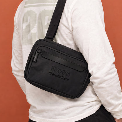 His Good Stuff's Autumn Catalog HGS26S4 Urban Bag, front closeup view, sleek, functional design with an adjustable shoulder strap and a secure zip closure, on a model against a flat Brandied Melon background. Black with black-thread-embroidered letters "HGS26S4" and HisGoodStuff.com underneath