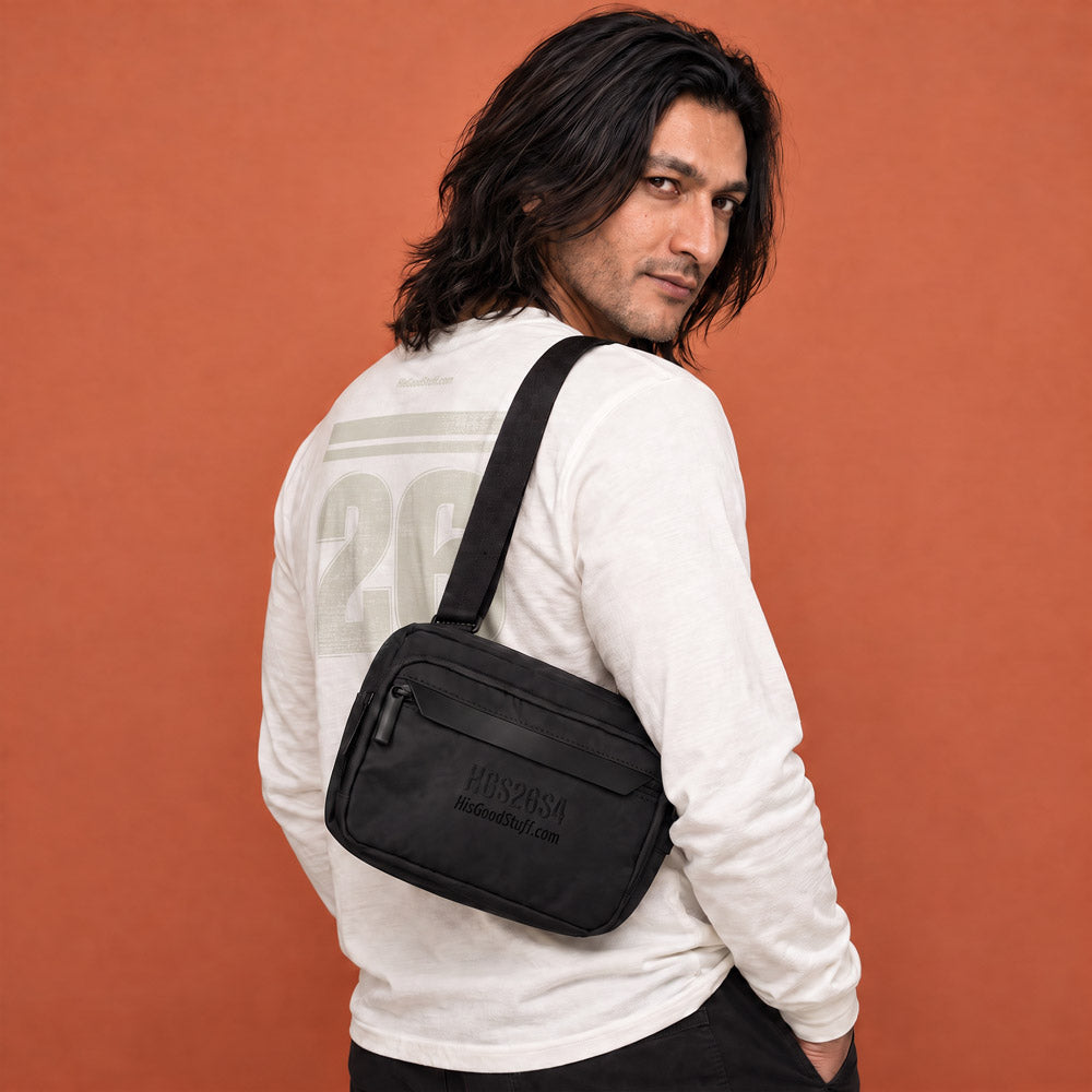 His Good Stuff's Autumn Catalog HGS26S4 Urban Bag, front view, sleek, functional design with an adjustable shoulder strap and a secure zip closure, on a model against a flat Brandied Melon background. Black with black-thread-embroidered letters "HGS26S4" and HisGoodStuff.com underneath
