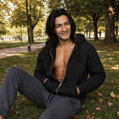 His Good Stuff's Autumn Catalog HGS26S4 Zip Hoodie, front, relaxed fit, black fleece, matching round drawcord with metal eyelets and tipped end, on a model sitting on the grass in a city park in Fall. Graphic displays in the top right a black embroidered HGS Fall thumbs-up icon with HisGoodStuff.com underneath