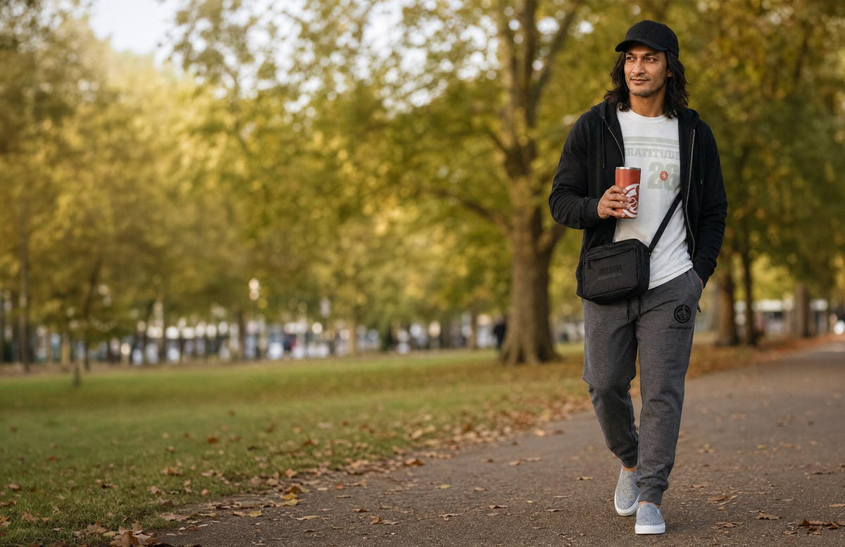 A man walking on a tree-lined path in a park, wearing a black zip-up hoodie, white graphic t-shirt, gray sweatpants, and a black cap, while holding a red cup.