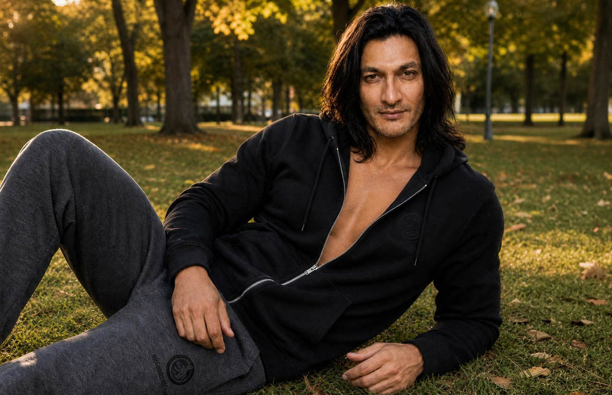 A man with long dark hair reclines on grass in a park, wearing a black zip-front hoodie with the front open and dark gray sweatpants. Trees and grass are visible in the background.
