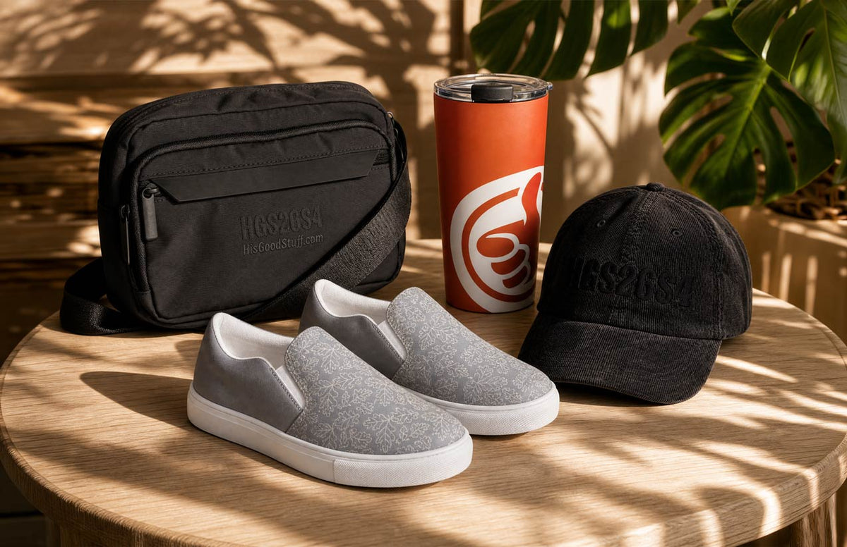 A styled product display for His Good Stuff featuring a black crossbody bag, gray slip-on sneakers with white soles, a black baseball cap, and an orange travel tumbler with a white logo, arranged on a round wooden table in the shade of leafy branches.